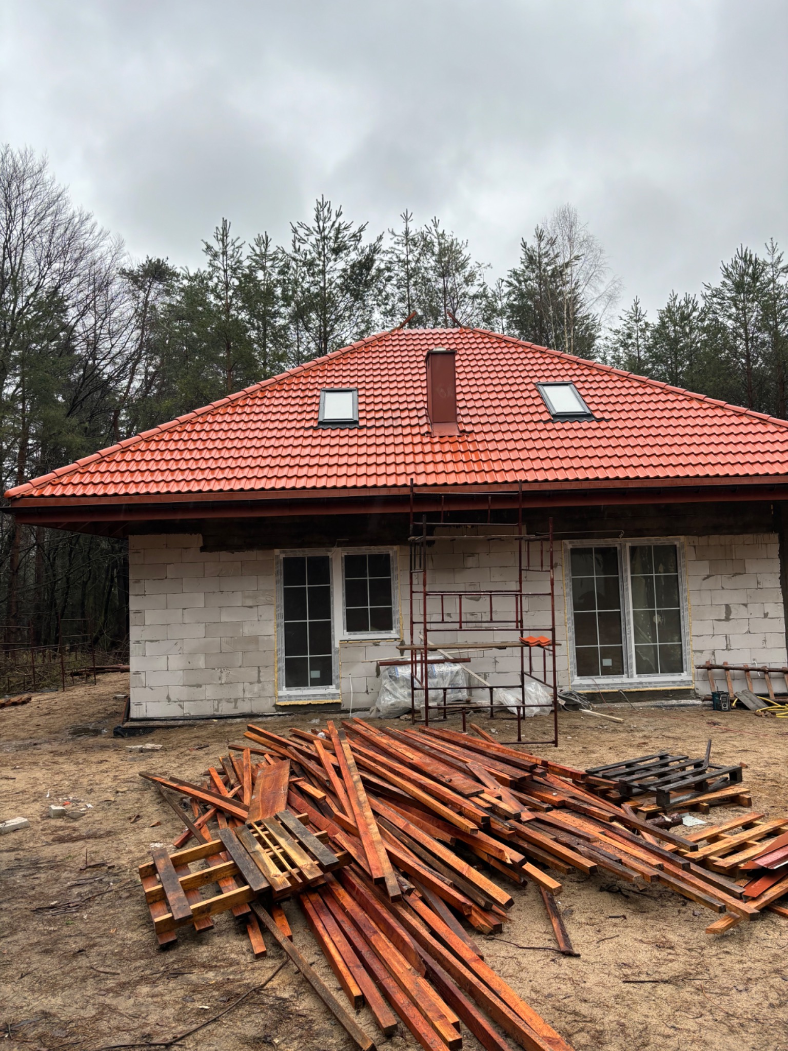 Dom w trakcie budowy z czerwonym dachem z dachówki ceramicznej, widoczne okna dachowe, ściany z bloczków, rusztowanie i sterta mokrego drewna na placu budowy w pochmurny dzień.