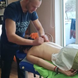 ADAM - Mężczyzna w niebieskiej koszulce Pierre Cardin wykonuje masaż pleców na leżącej kobiecie na niebiesko-zielonym stole do masażu w pokoju z oknem.