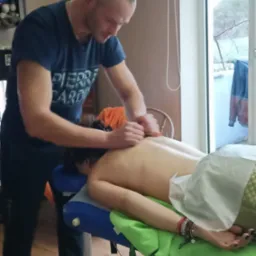 Mężczyzna w niebieskiej koszulce Pierre Cardin wykonuje masaż pleców na leżącej kobiecie na niebiesko-zielonym stole do masażu w pokoju z oknem.