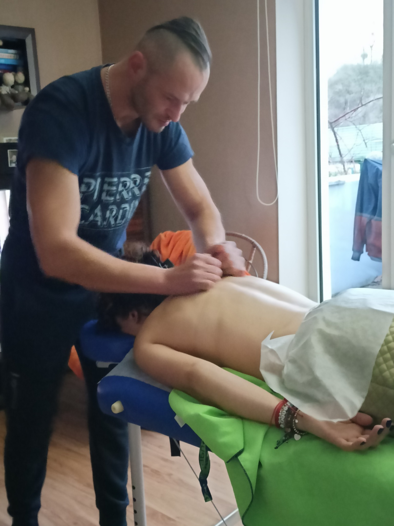 Mężczyzna w niebieskiej koszulce Pierre Cardin wykonuje masaż pleców na leżącej kobiecie na niebiesko-zielonym stole do masażu w pokoju z oknem.