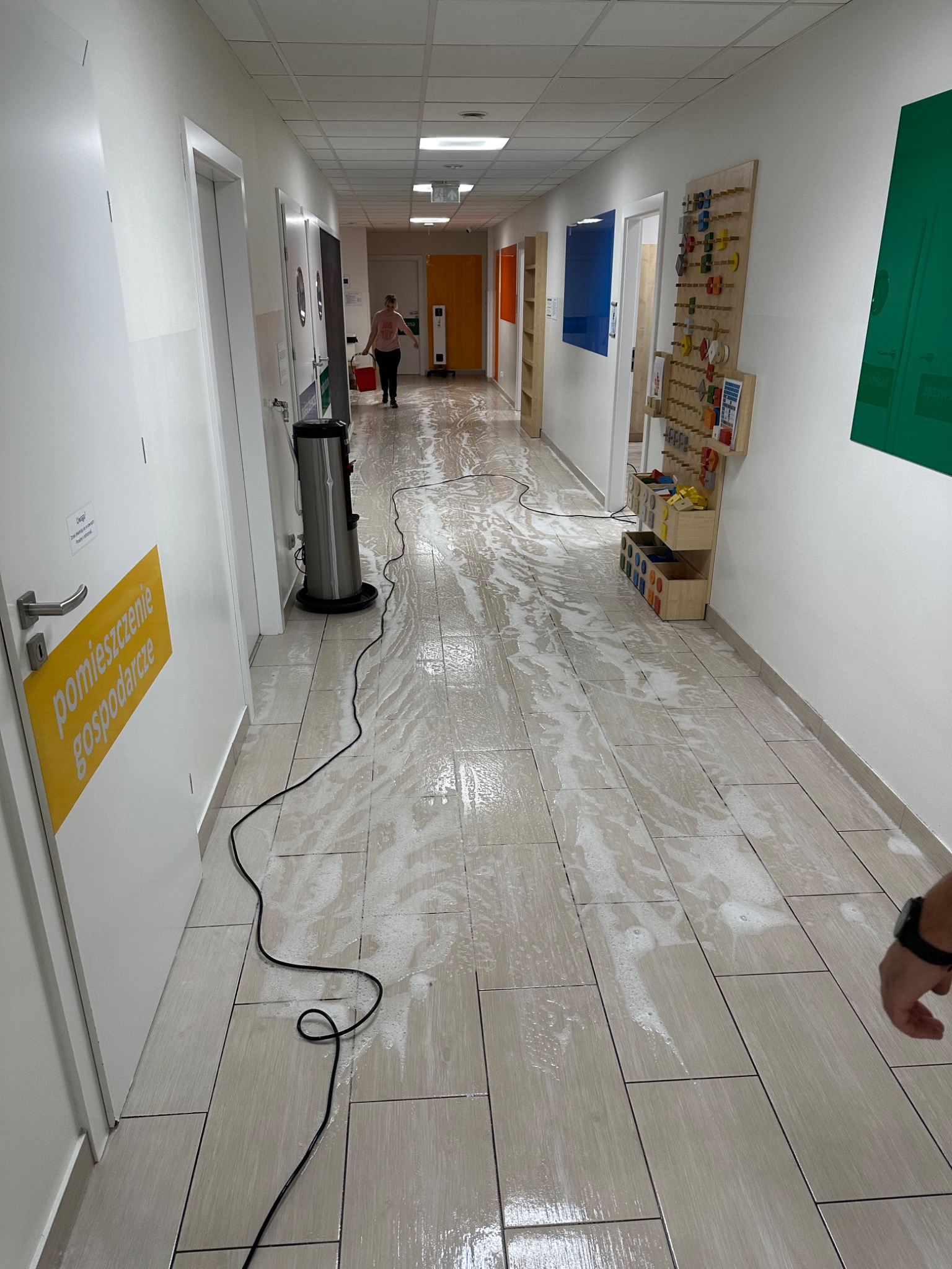 Korytarz w trakcie czyszczenia: podłoga pokryta pianą, widoczny odkurzacz piorący i osoba z wiadrem w tle, żółty napis 'pomieszczenie gospodarcze' na drzwiach po lewej.