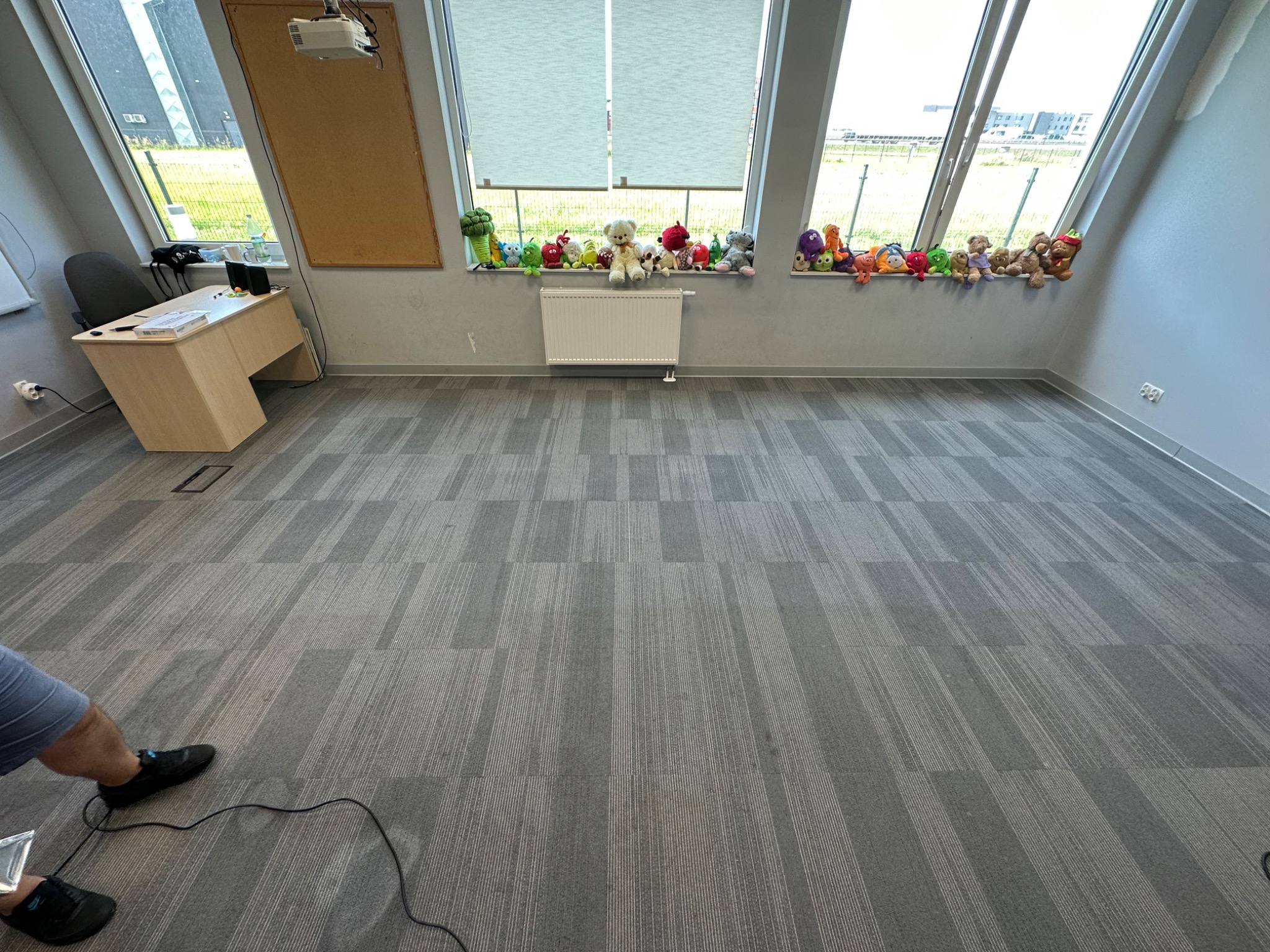 Przestronne biuro z szarym dywanem w geometryczne wzory, biurkiem przy oknie i kolekcją pluszowych zabawek na parapecie.