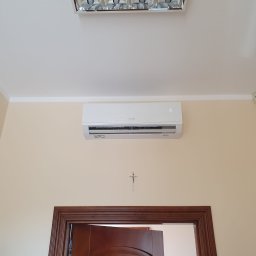 AEROKLIM Oleksandr Bashov - Biały klimatyzator ścienny zamontowany nad drewnianą futryną drzwi w pokoju o kremowych ścianach, pod sufitem widoczna lampa rastrowa, a na ścianie mały krzyżyk.