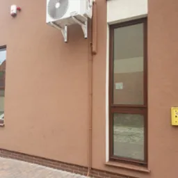 Zewnętrzna jednostka klimatyzacji Sinclair zamontowana na brązowej elewacji budynku mieszkalnego, widoczne brązowe rury odprowadzające skropliny, okno z brązową ramą i chodnik z kostki brukowej.