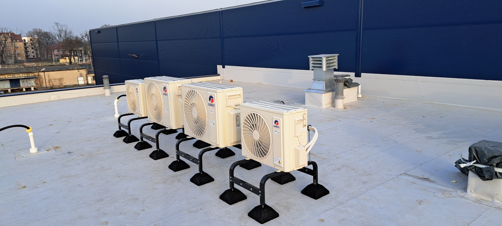 Rząd kremowych jednostek zewnętrznych klimatyzacji marki Gree, zamontowanych na czarnych metalowych podstawach na płaskim dachu budynku z niebieską fasadą.