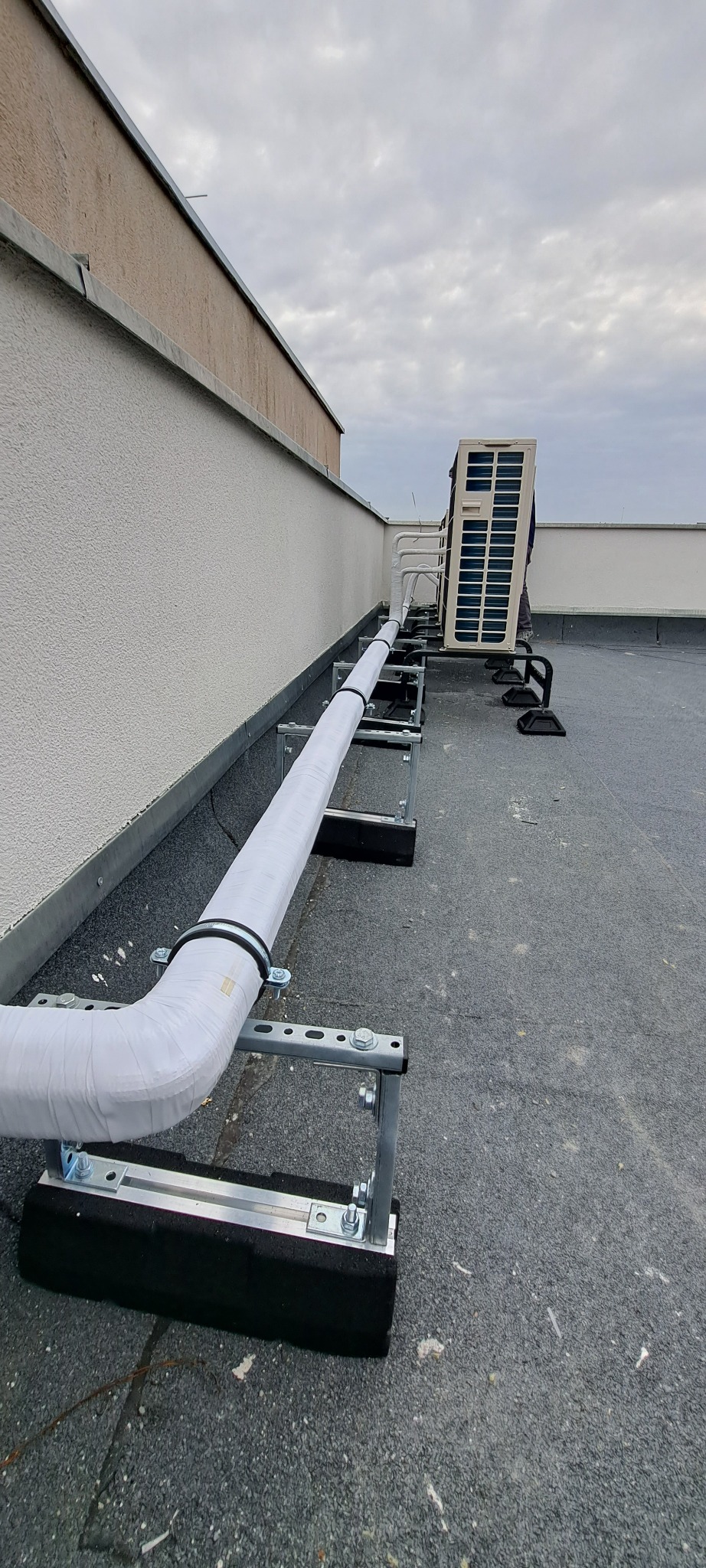 Instalacja klimatyzacji na dachu budynku: rury w izolacji termicznej prowadzone na metalowych wspornikach, jednostka zewnętrzna klimatyzatora w tle.