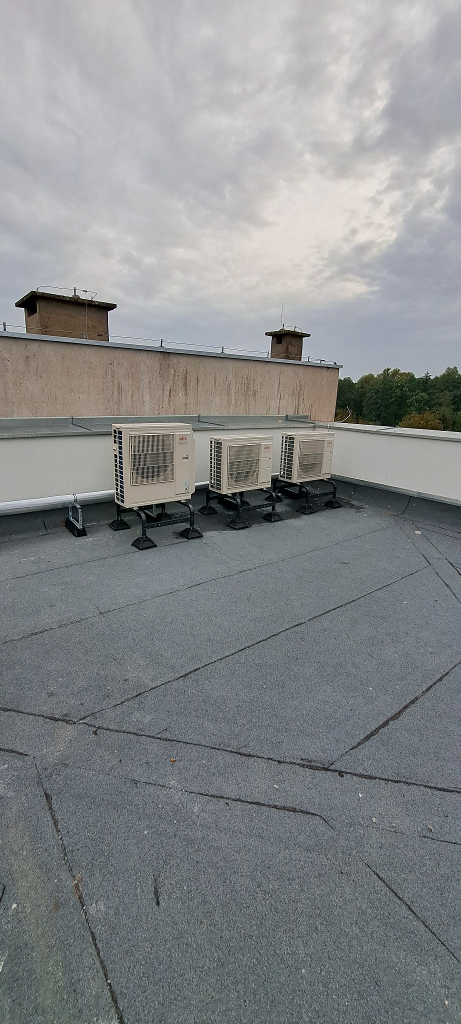 Trzy zewnętrzne jednostki klimatyzacji Fujitsu zamontowane na czarnych wspornikach na płaskim dachu budynku, widoczne kominy i pochmurne niebo.