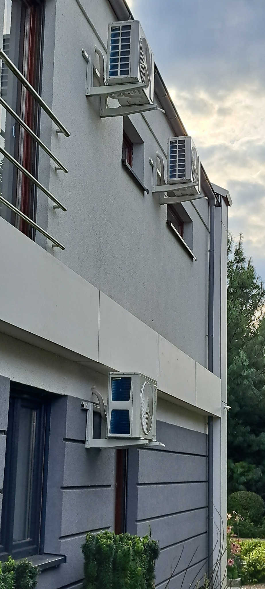 Trzy klimatyzatory zamontowane na elewacji domu z widocznymi oknami i fragmentem ogrodu w tle, uchwycone w pochmurny dzień.