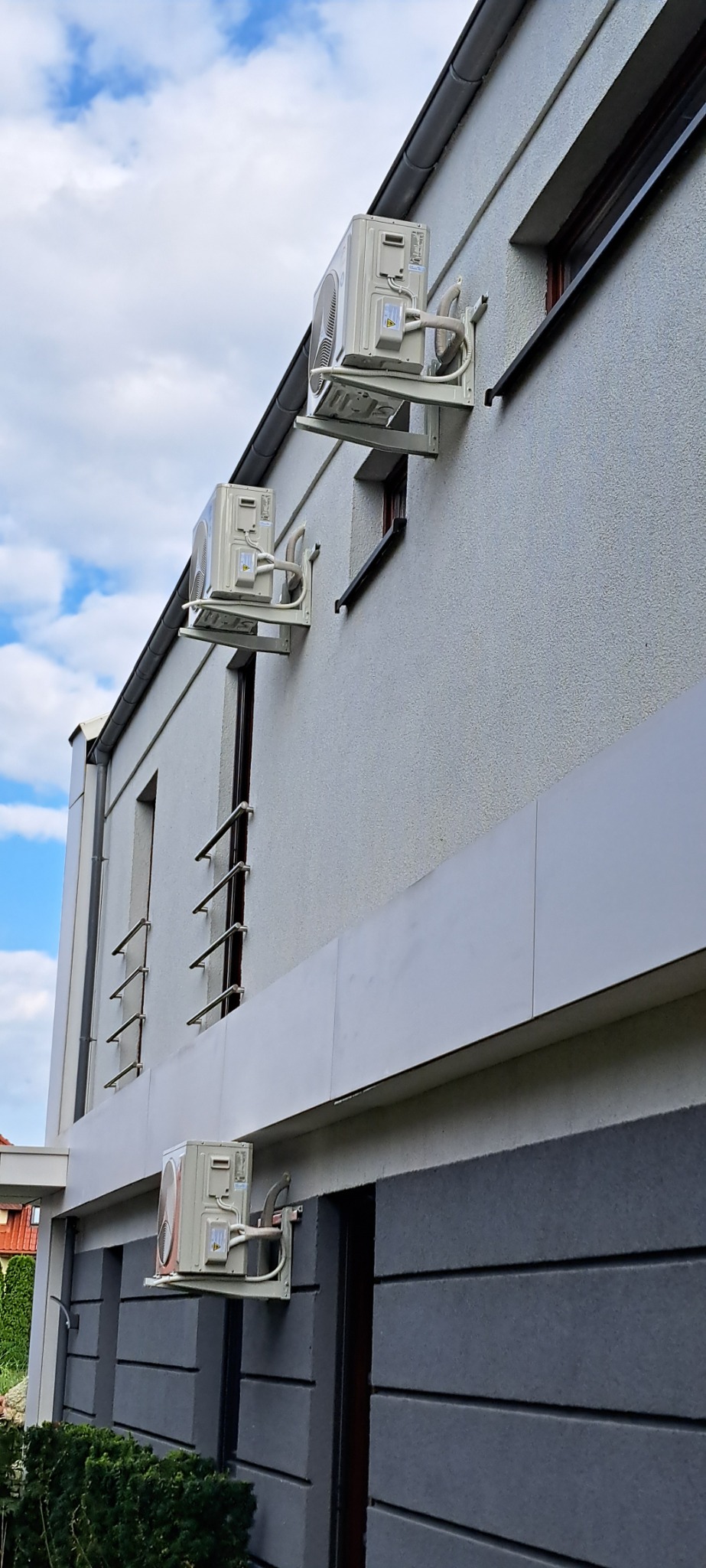 Trzy klimatyzatory zamontowane na elewacji budynku mieszkalnego, widoczne rury odprowadzające skropliny, ściana pomalowana na szaro z elementami dekoracyjnymi.