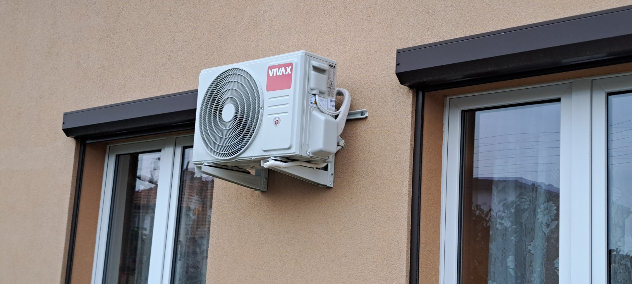 Zewnętrzna jednostka klimatyzacji Vivax zamontowana na elewacji domu między dwoma oknami z roletami zewnętrznymi.