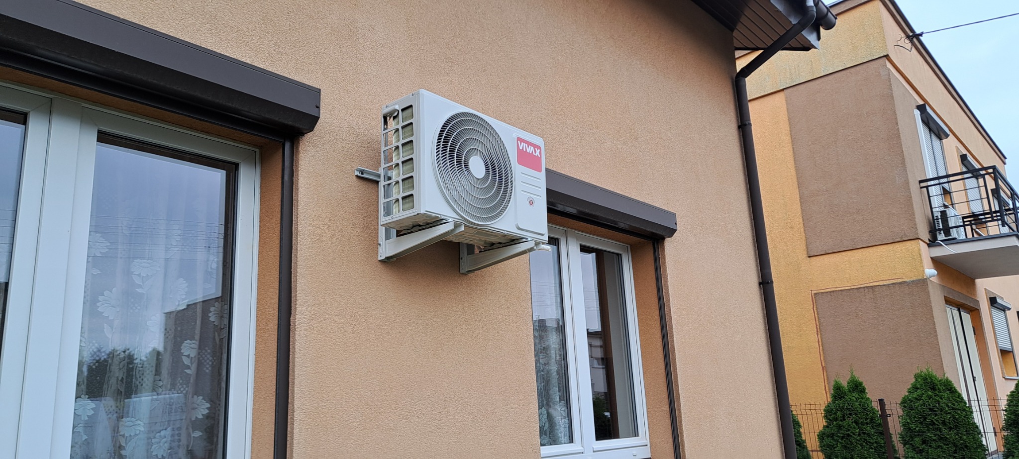 Zewnętrzna jednostka klimatyzacji VIVAX zamontowana na elewacji budynku w Poznaniu, widoczna z boku, z brązowymi roletami okiennymi w tle.