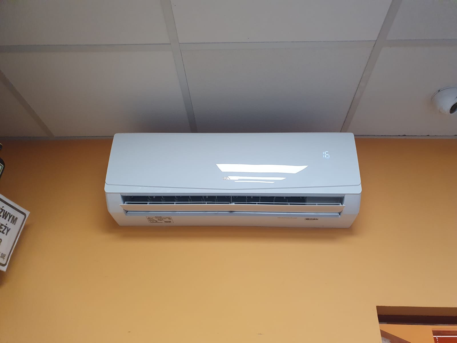 Biała klimatyzacja ścienna z wyświetlaczem temperatury 16 stopni, zamontowana na pomarańczowej ścianie pod sufitem kasetonowym.