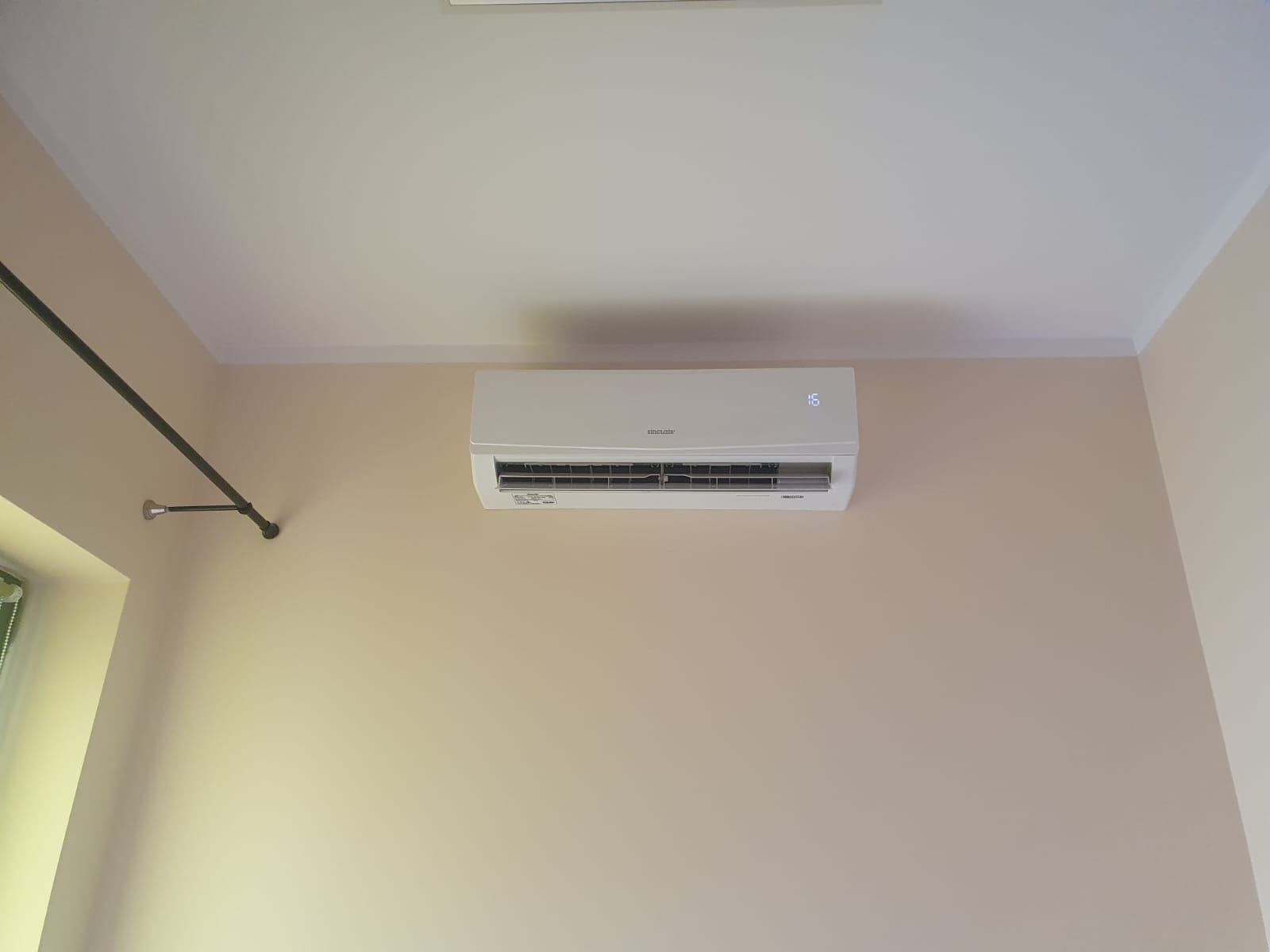 Biała jednostka wewnętrzna klimatyzacji marki Sinclair zamontowana na beżowej ścianie w pomieszczeniu. Na wyświetlaczu widoczna temperatura 16 stopni Celsjusza. Po lewej stronie widoczny fragment...
