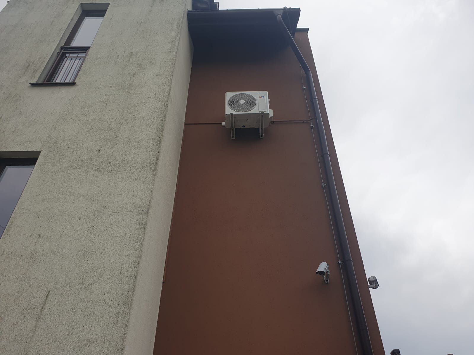 Zewnętrzna jednostka klimatyzacji zamontowana na brązowej ścianie budynku mieszkalnego, widoczne rury odprowadzające skropliny i kamery monitoringu.