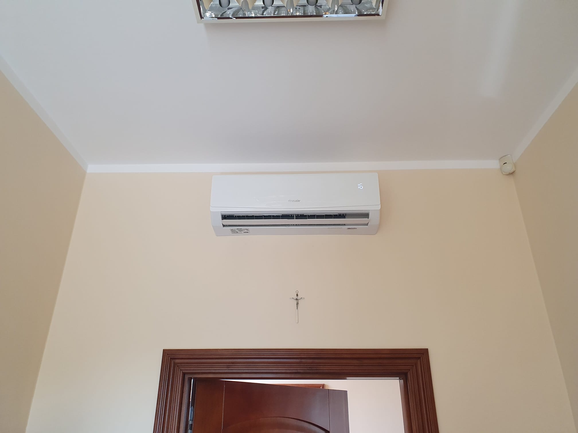 Biały klimatyzator ścienny zamontowany nad drewnianą futryną drzwi w pokoju o kremowych ścianach, pod sufitem widoczna lampa rastrowa, a na ścianie mały krzyżyk.