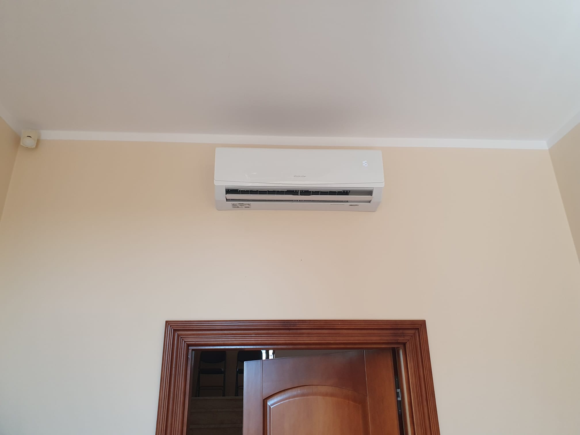 Biały klimatyzator ścienny zamontowany nad drewnianymi drzwiami w jasnym pomieszczeniu, widoczny wyświetlacz temperatury.