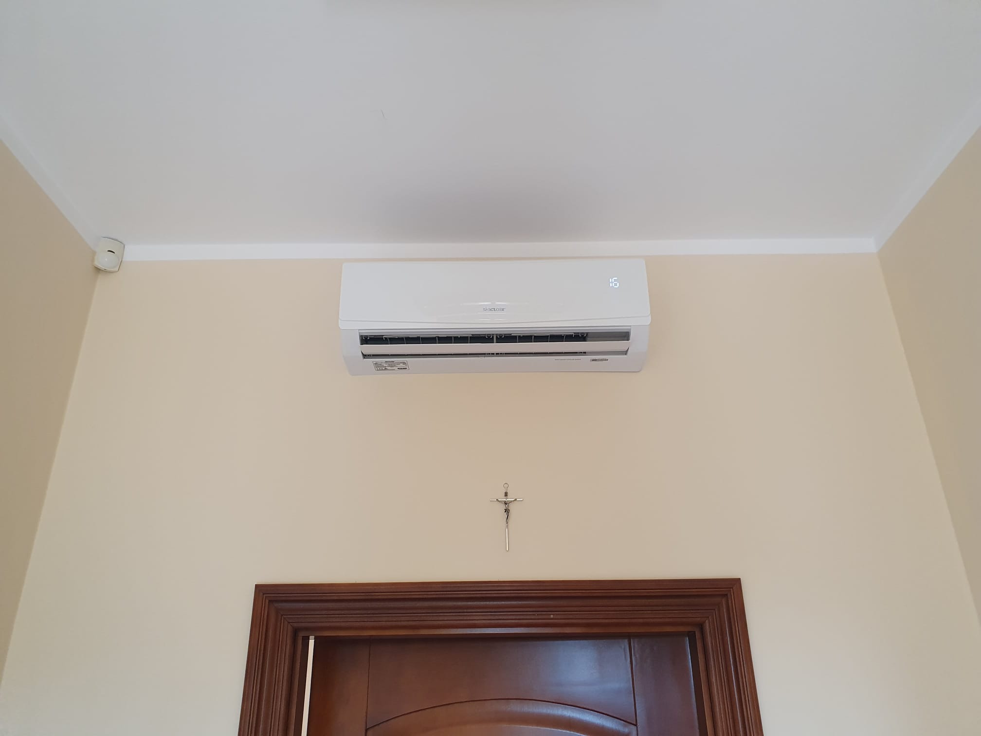 Biały klimatyzator ścienny zamontowany w pokoju z beżowymi ścianami, widoczny krzyżyk dekoracyjny nad ciemnobrązowymi drzwiami.