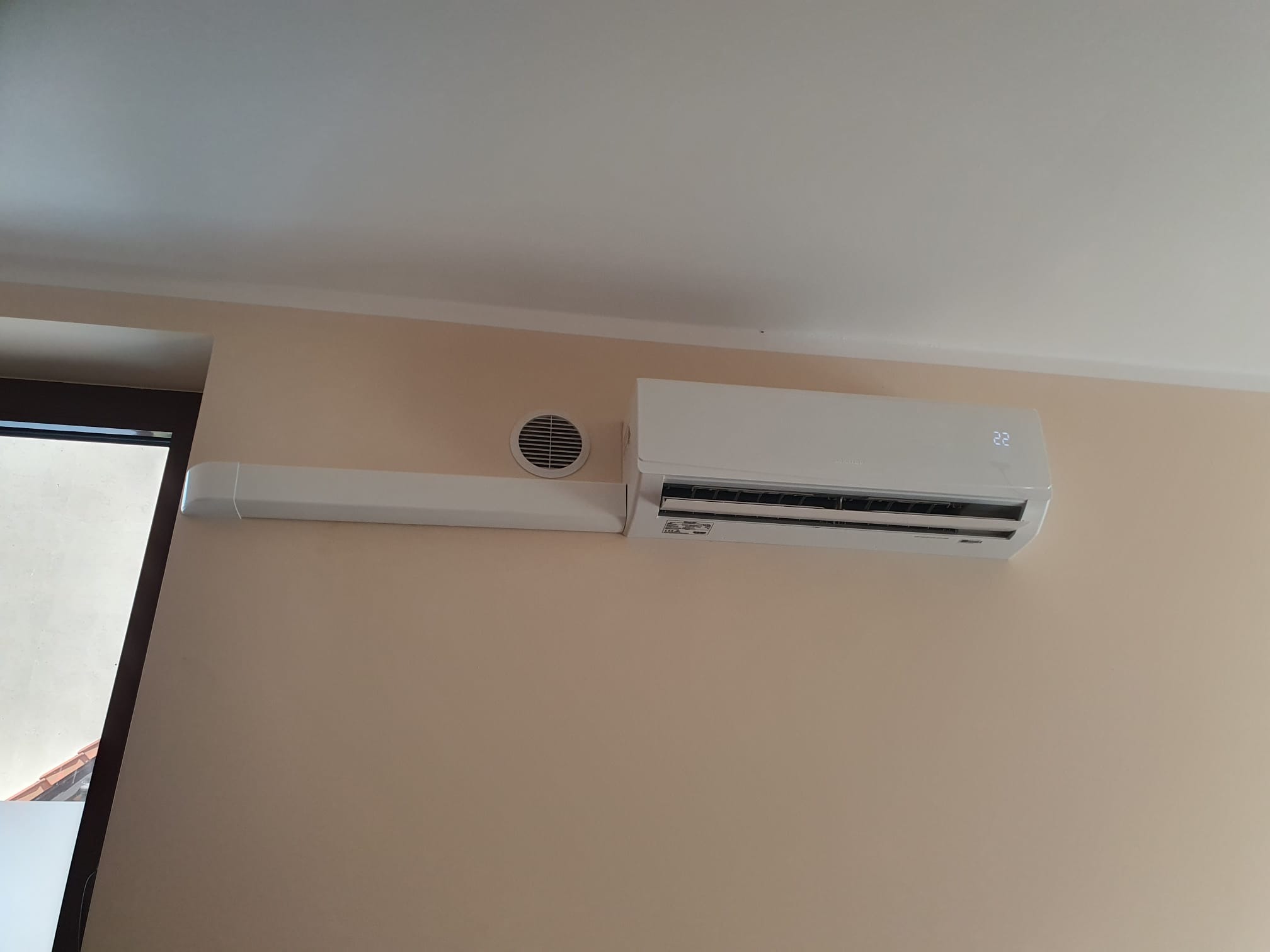 Biała klimatyzacja ścienna z widocznym wyświetlaczem temperatury (22 stopnie Celsjusza) i maskownicą rur, zamontowana na beżowej ścianie w pomieszczeniu z wentylacją.