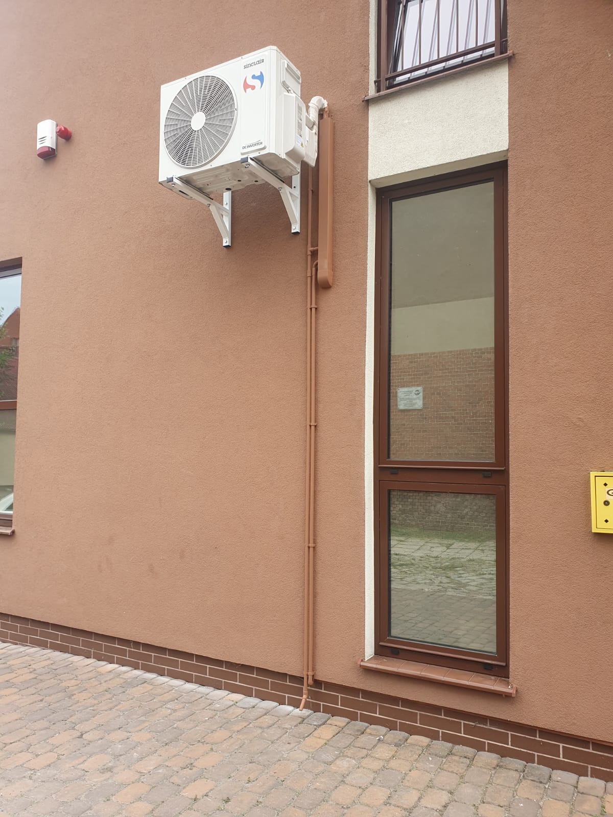 Zewnętrzna jednostka klimatyzacji Sinclair zamontowana na brązowej elewacji budynku mieszkalnego, widoczne brązowe rury odprowadzające skropliny, okno z brązową ramą i chodnik z kostki brukowej.