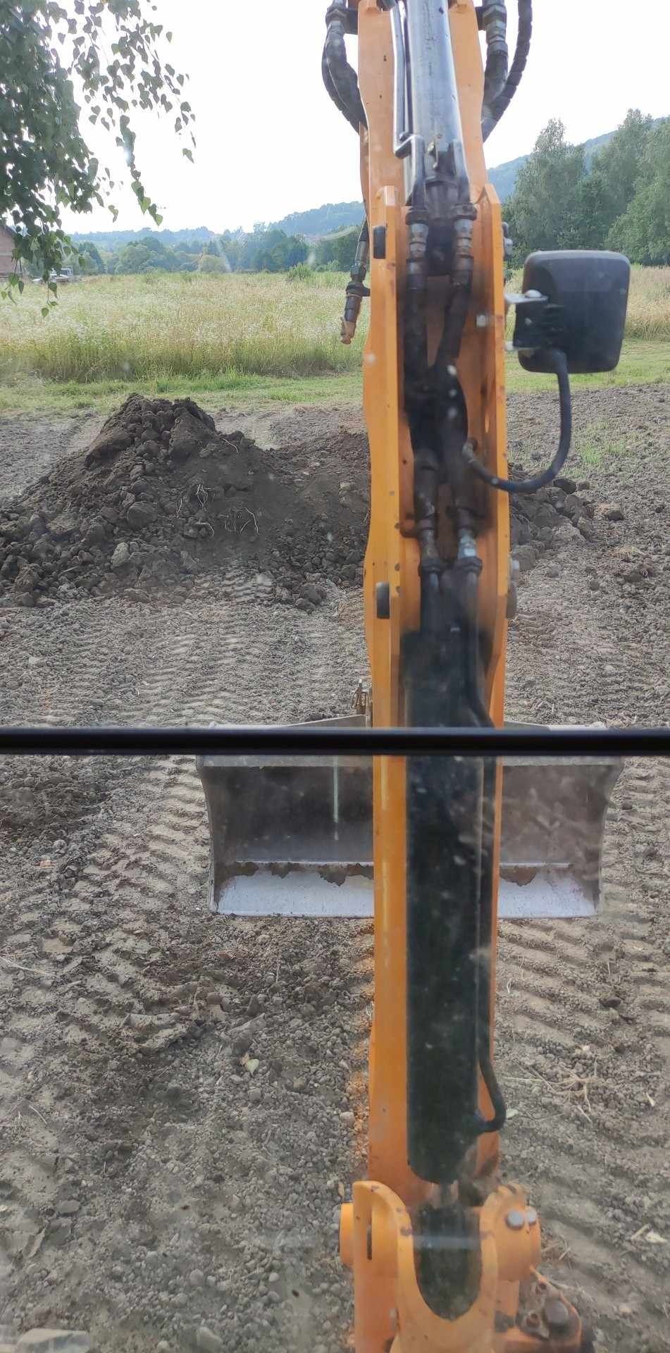 Widok z kabiny koparki na wykopany grunt i stertę ziemi na tle zielonego pola i drzew