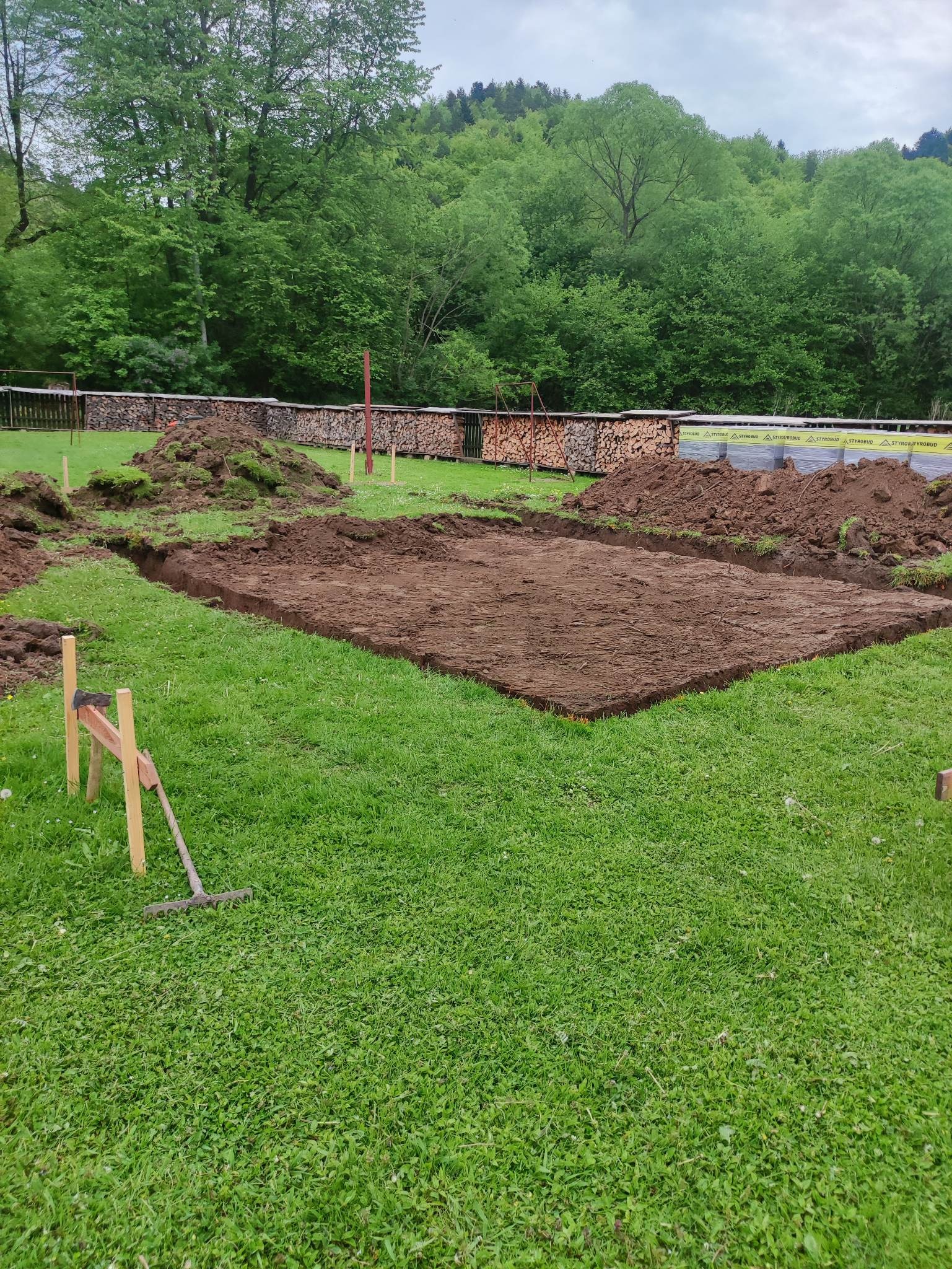 Przygotowany prostokątny wykop pod fundamenty na trawiastym terenie, z hałdami ziemi po bokach i drewnianymi stemplami w narożnikach, w tle las i składowane drewno.