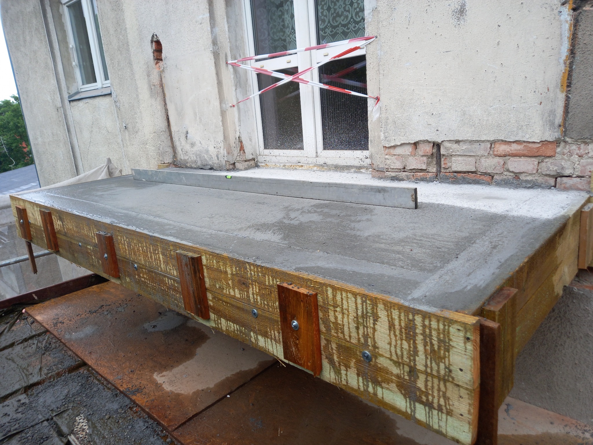 Wylewanie balkonu: świeży beton w drewnianym szalunku, poziomica na wierzchu, zabezpieczone okno w tle.