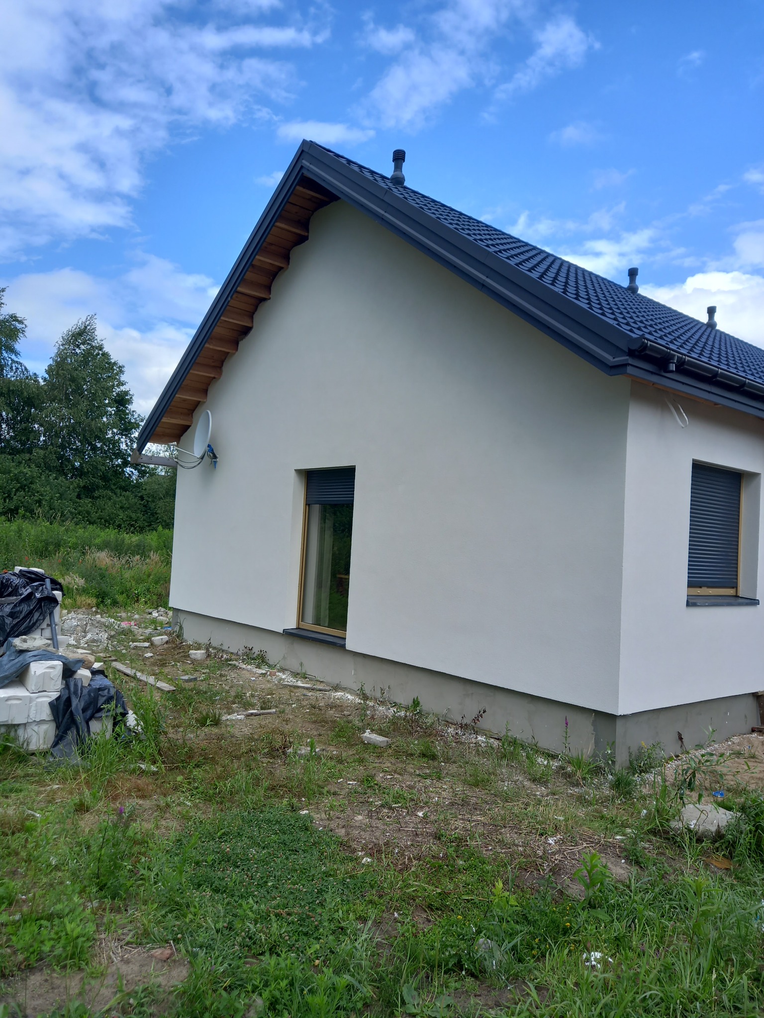 Nowo otynkowany, parterowy dom z ciemną dachówką i roletami zewnętrznymi, widok z boku na tle błękitnego nieba i zieleni, z widoczną anteną satelitarną i stertą materiałów budowlanych.