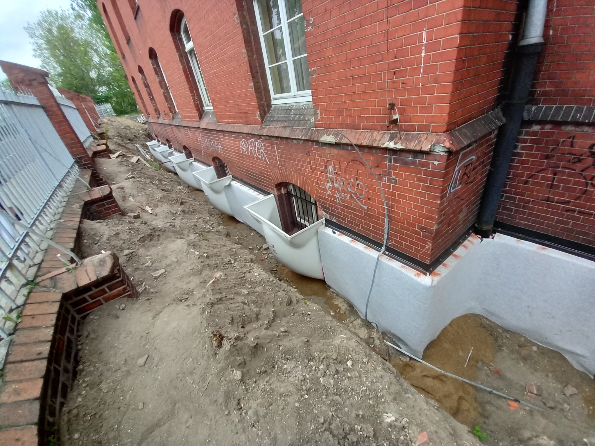 Ceglany budynek z odkrytymi fundamentami zabezpieczonymi białą folią izolacyjną i systemem wentylacji piwnic w postaci białych kominków wentylacyjnych.