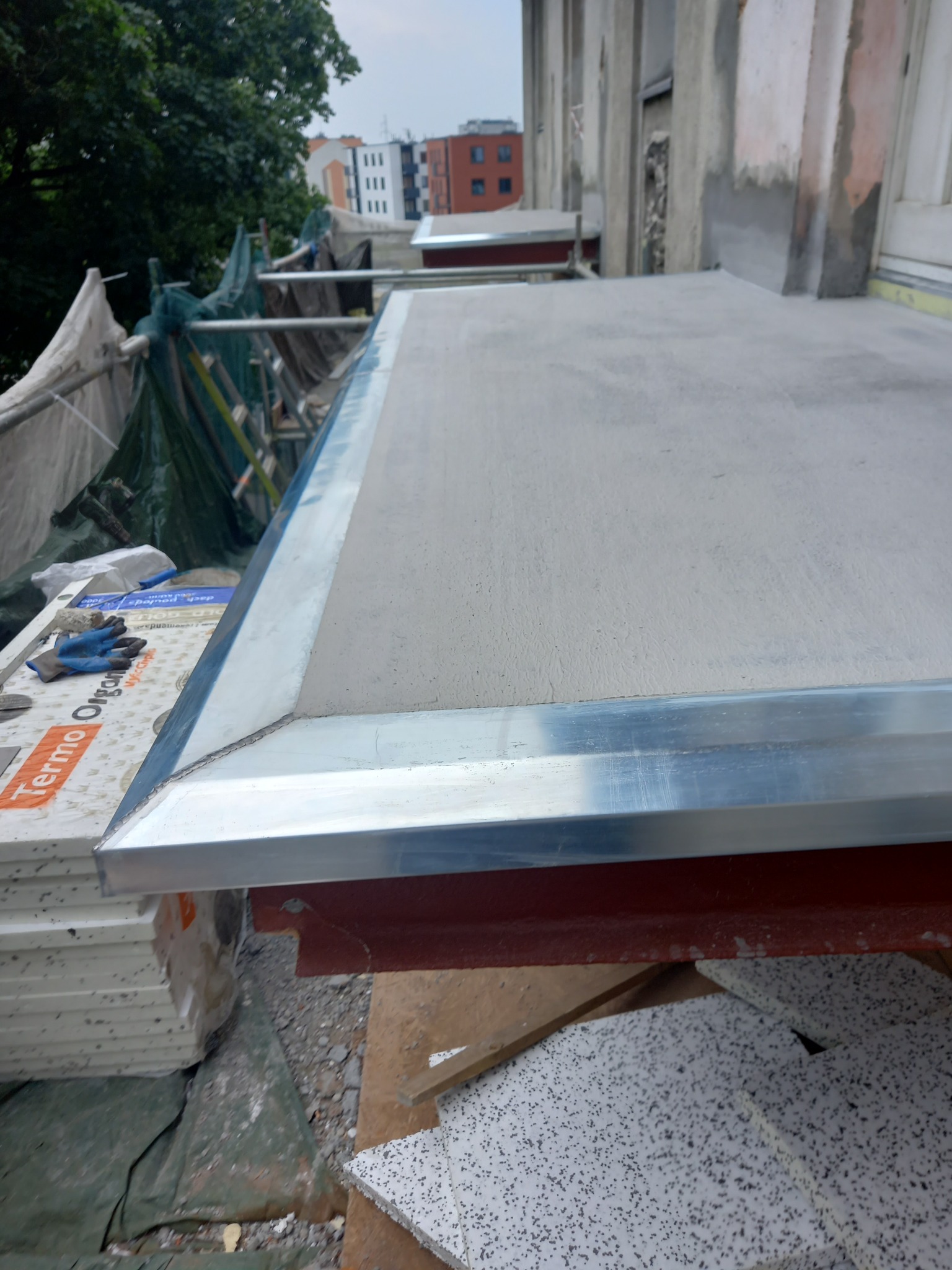 Ocieplony balkon z aluminiowym wykończeniem krawędzi, widoczne płyty styropianowe Terma Organika i robocze rękawice, rusztowanie w tle.