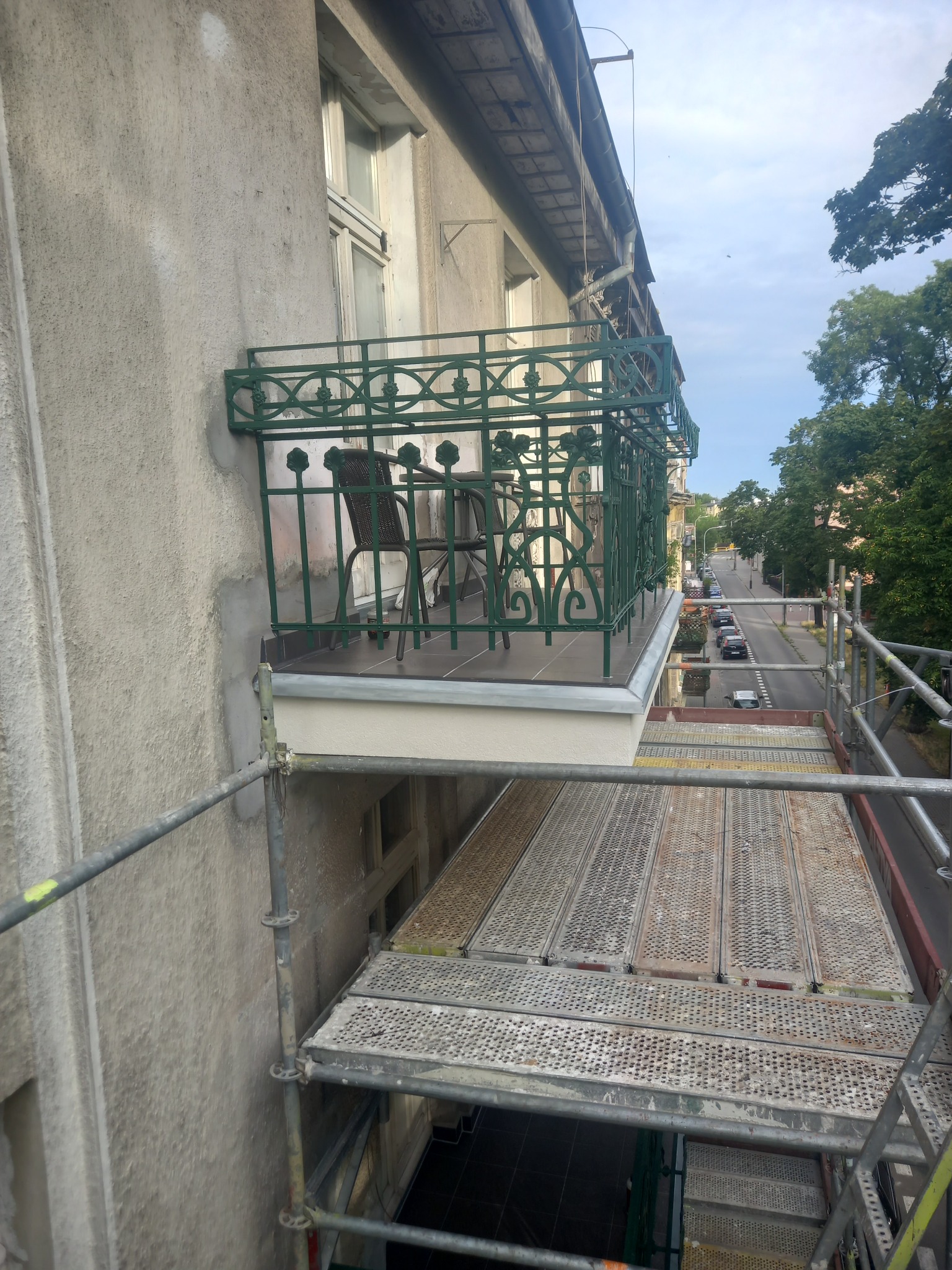 Renowacja balkonu z zieloną, kutą balustradą, widok z rusztowania na ulicę i budynek z odświeżoną elewacją.