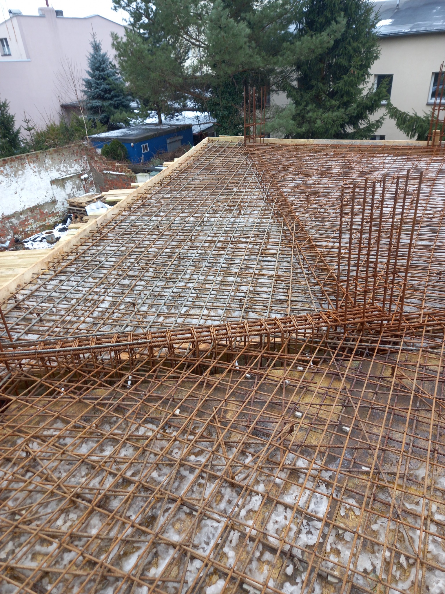 Zbrojenie stropu z siatki stalowej, widoczne drewniane szalunki i otoczenie z budynkami i drzewami w tle.
