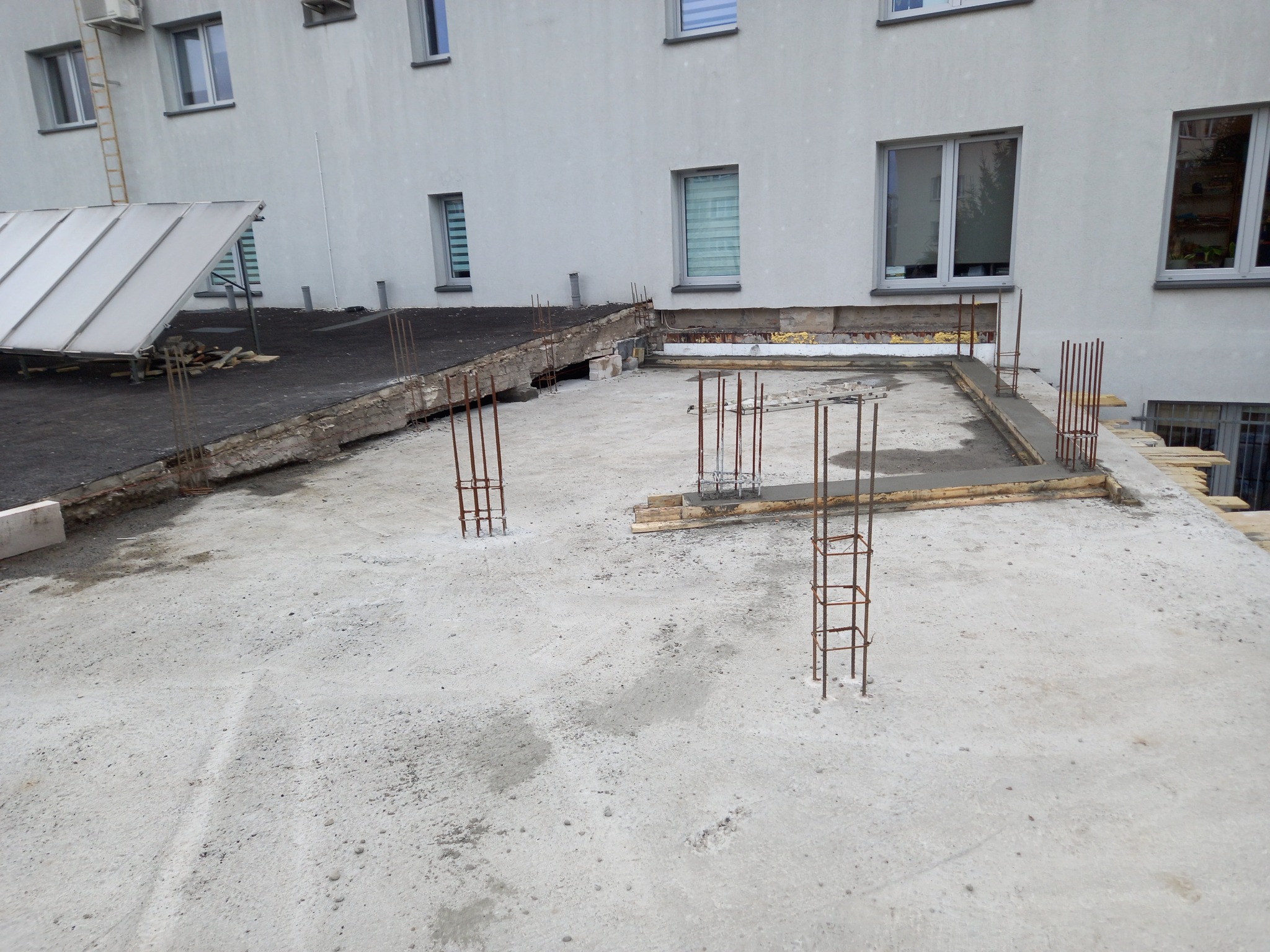 Betonowy strop w trakcie budowy z wystającymi prętami zbrojeniowymi, widoczne szalunki i fragment budynku w tle.