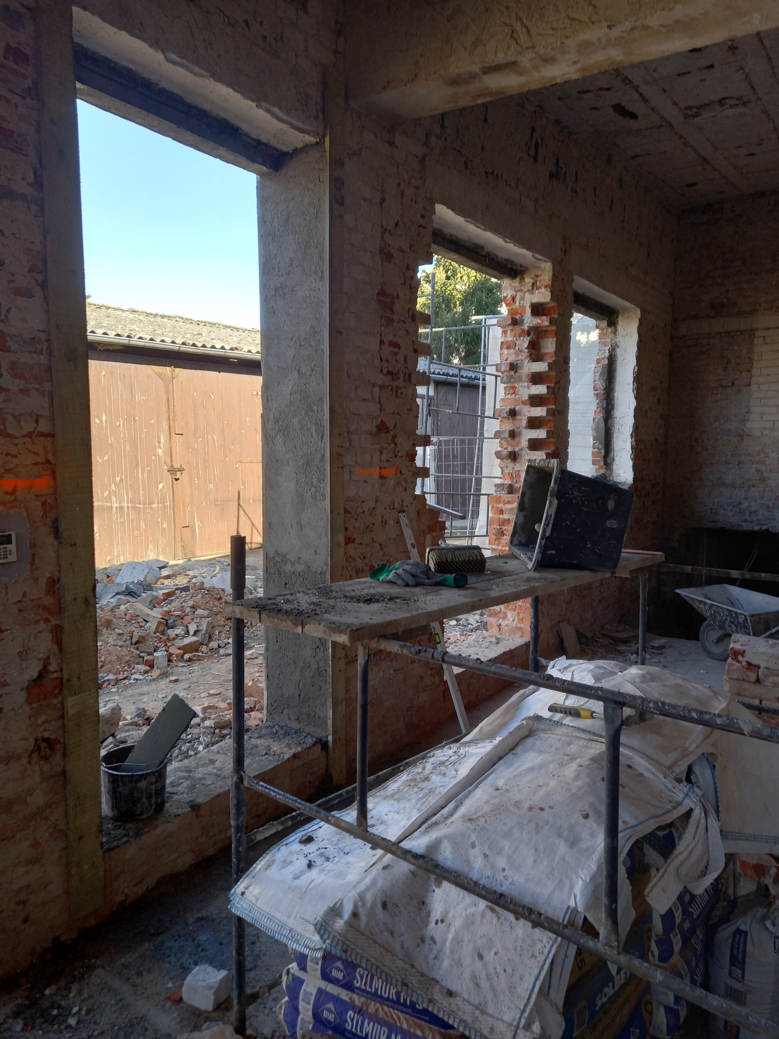 Wnętrze budynku w trakcie remontu, widoczne fragmenty ceglanych ścian, prowizoryczne rusztowanie z narzędziami, worki z materiałami budowlanymi oraz gruz na zewnątrz przez otwory okienne.