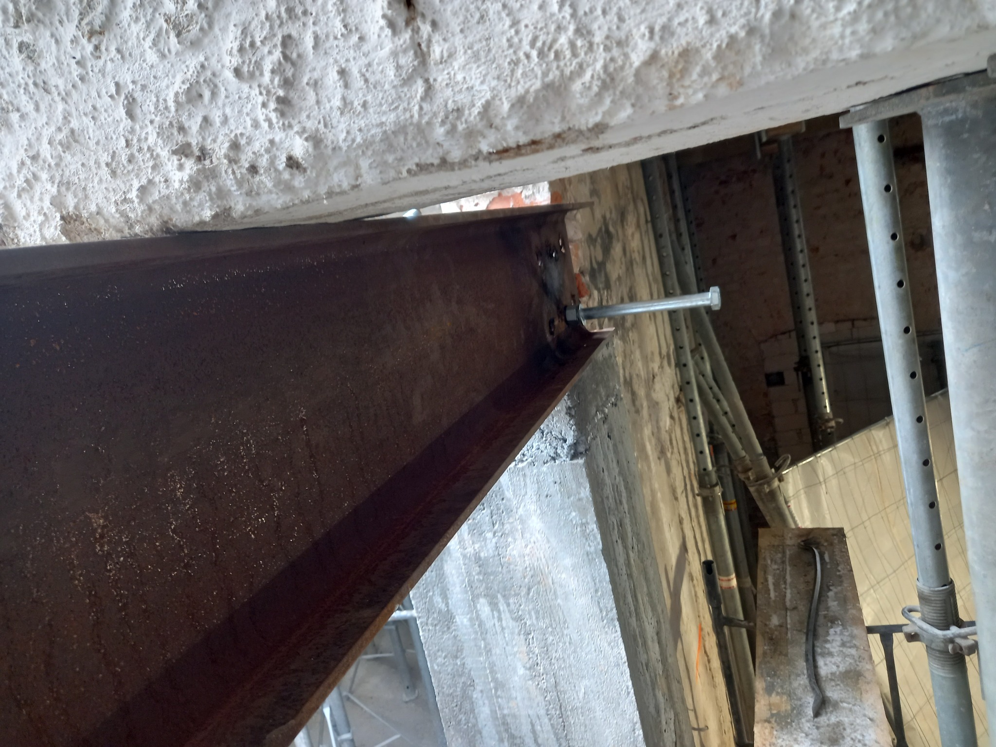 Stalowa belka mocowana do sufitu za pomocą śruby, widoczne podpory montażowe i fragment muru z nierówną fakturą.