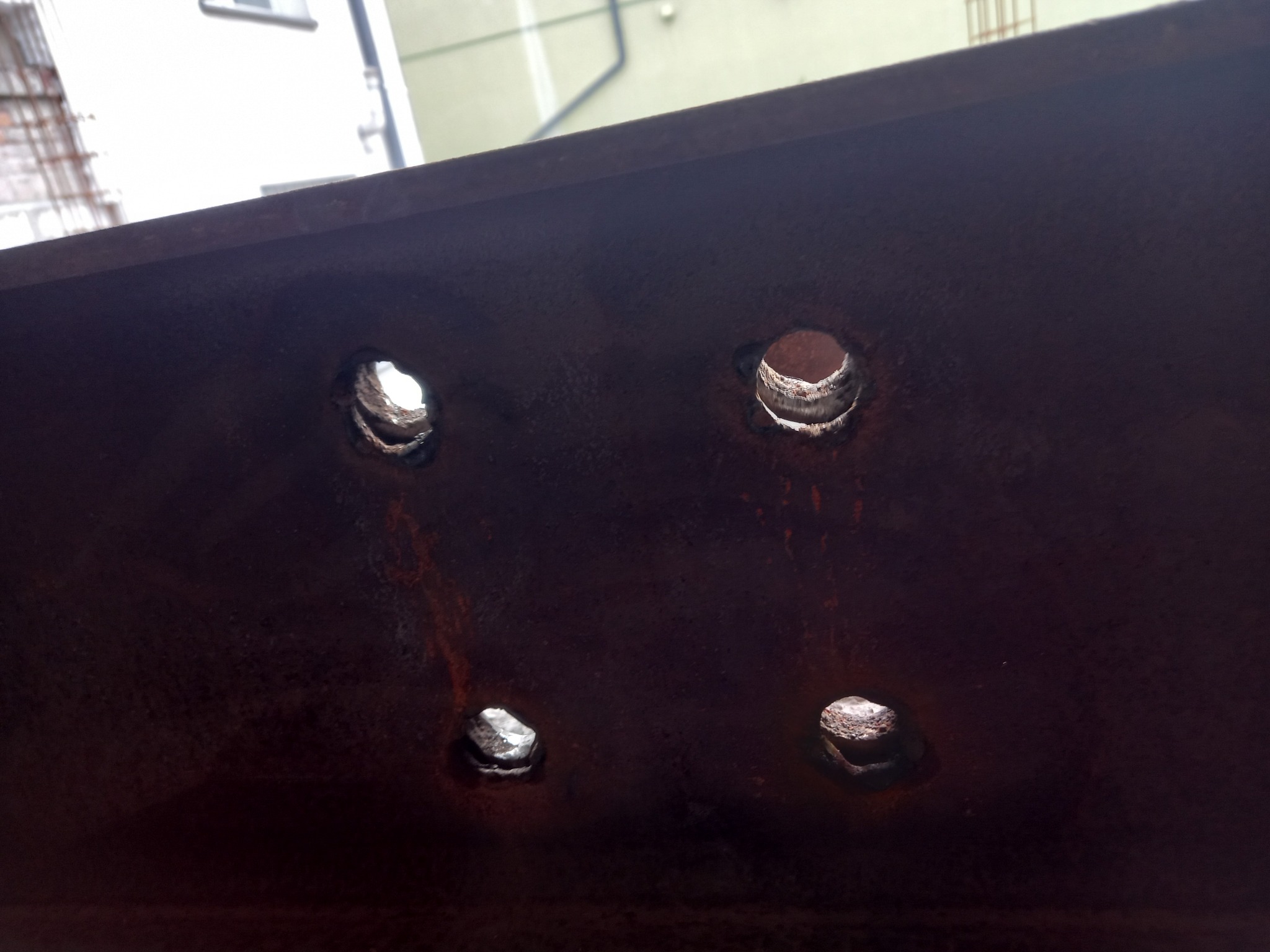 Metalowa belka z czterema wywierconymi otworami, widoczne ślady rdzy i obróbki metalu.