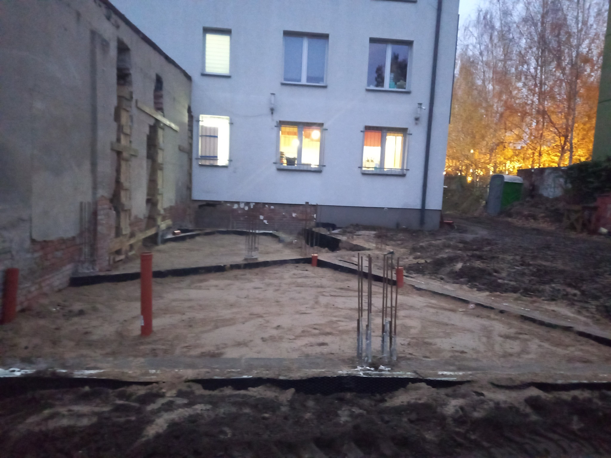 Przygotowanie terenu pod fundamenty nowego budynku, widoczne wystające pręty zbrojeniowe, rury kanalizacyjne i częściowo wykopane fundamenty na tle istniejącego budynku mieszkalnego i starego...