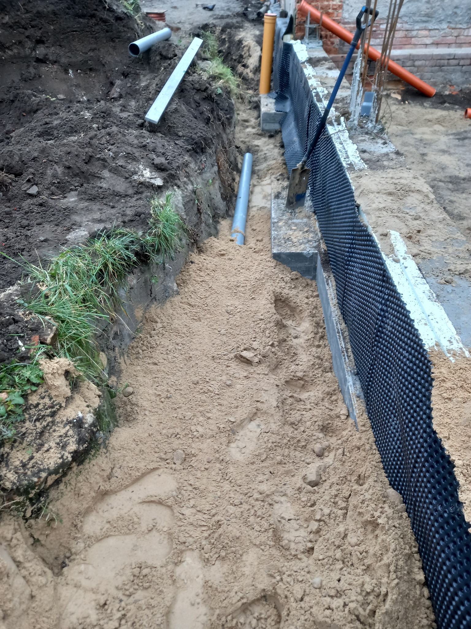 Wykop fundamentowy z izolacją przeciwwilgociową z folii kubełkowej, zasypywany piaskiem, widoczne rury kanalizacyjne i szpadel.