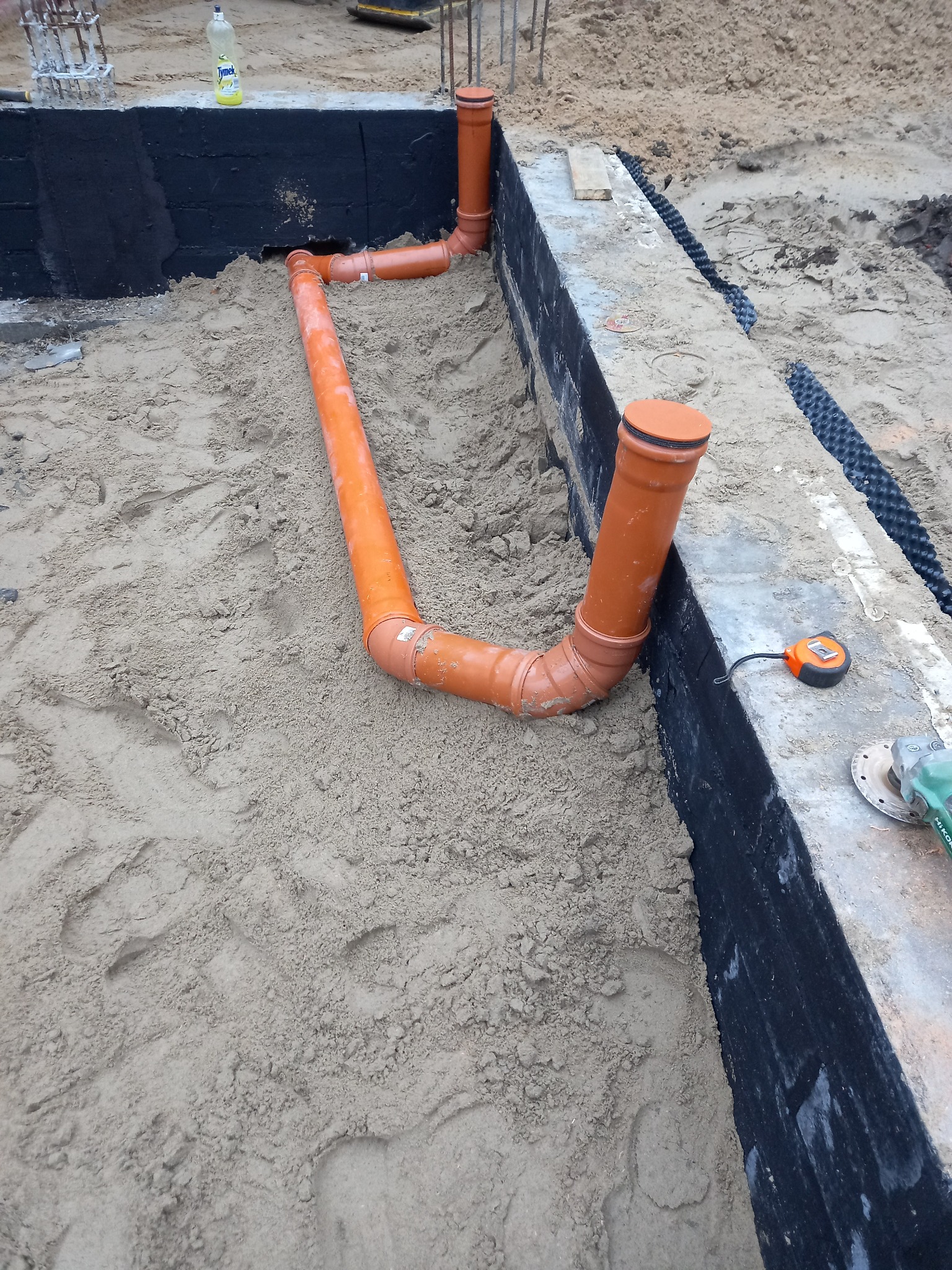 Pomarańczowe rury kanalizacyjne ułożone w wykopie fundamentowym, zabezpieczonym czarną izolacją, z widoczną taśmą mierniczą i szlifierką kątową na betonowej ławie.