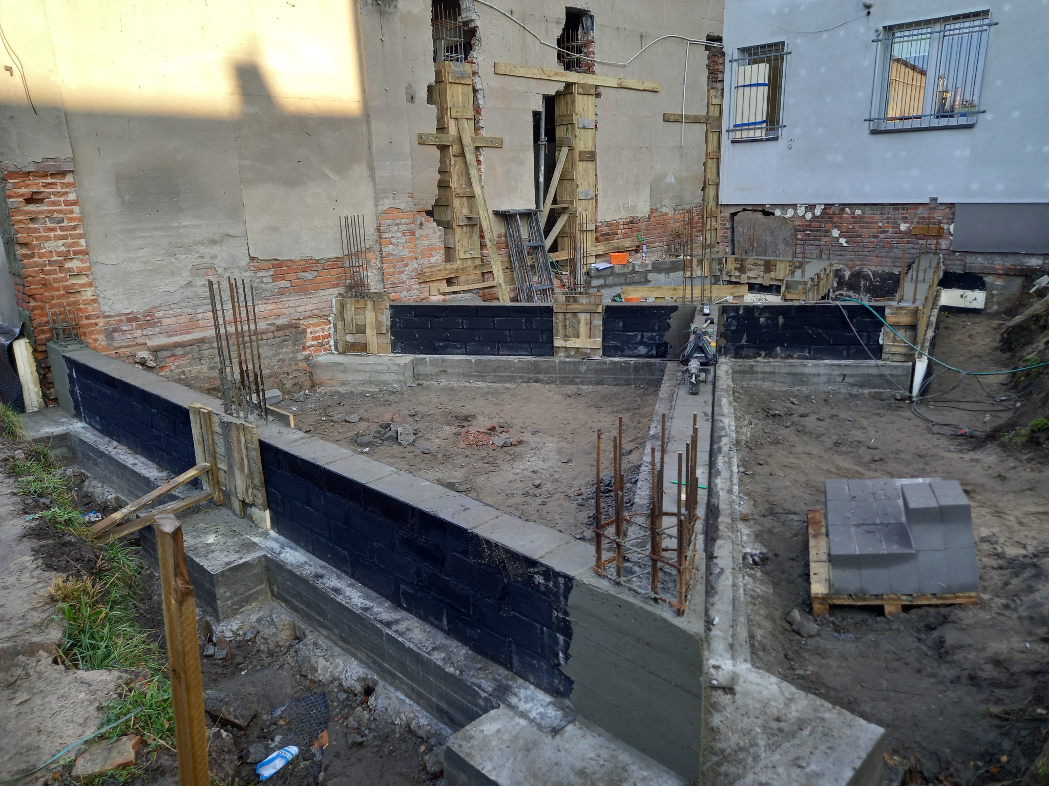 Fundamenty budynku w trakcie budowy, zabezpieczone czarną izolacją, z widocznymi prętami zbrojeniowymi i fragmentami deskowania. W tle ściana budynku z cegły i betonu, z oknami zabezpieczonymi...