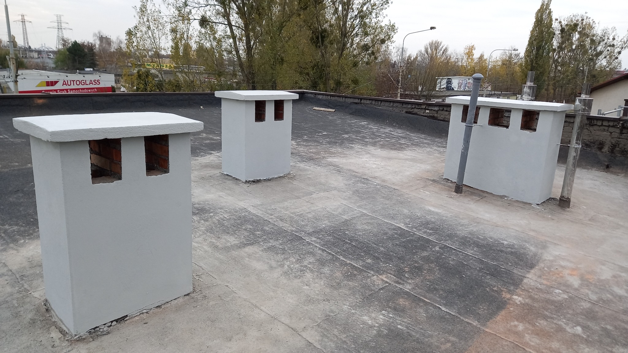 Trzy odnowione kominy wentylacyjne z ceglanymi otworami, pomalowane na szaro, na płaskim dachu pokrytym papą, w tle budynki i drzewa.