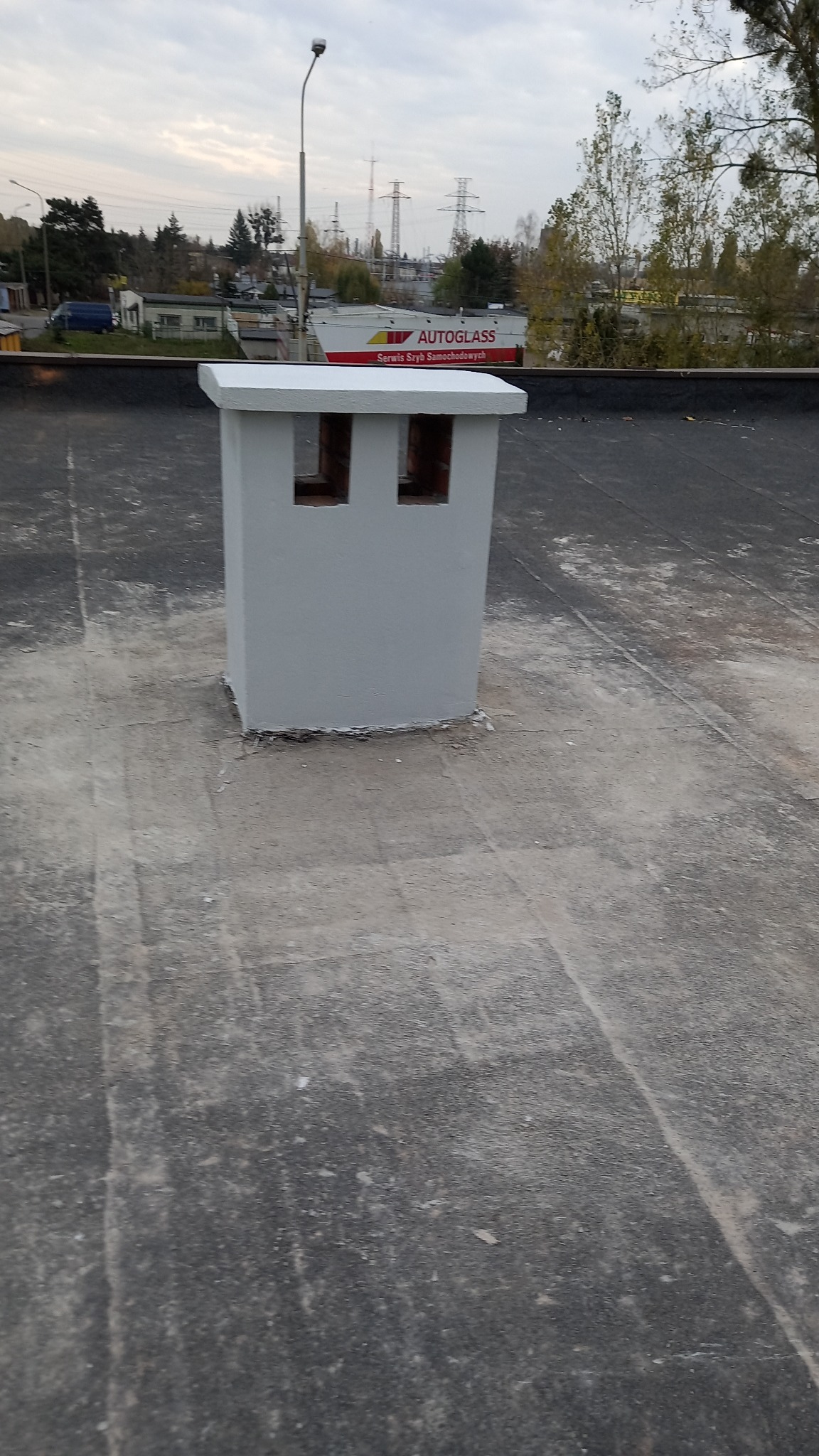 Szary komin wentylacyjny z dwoma otworami na płaskim, bitumicznym dachu budynku. W tle widoczny budynek z reklamą Autoglass i słup oświetleniowy.