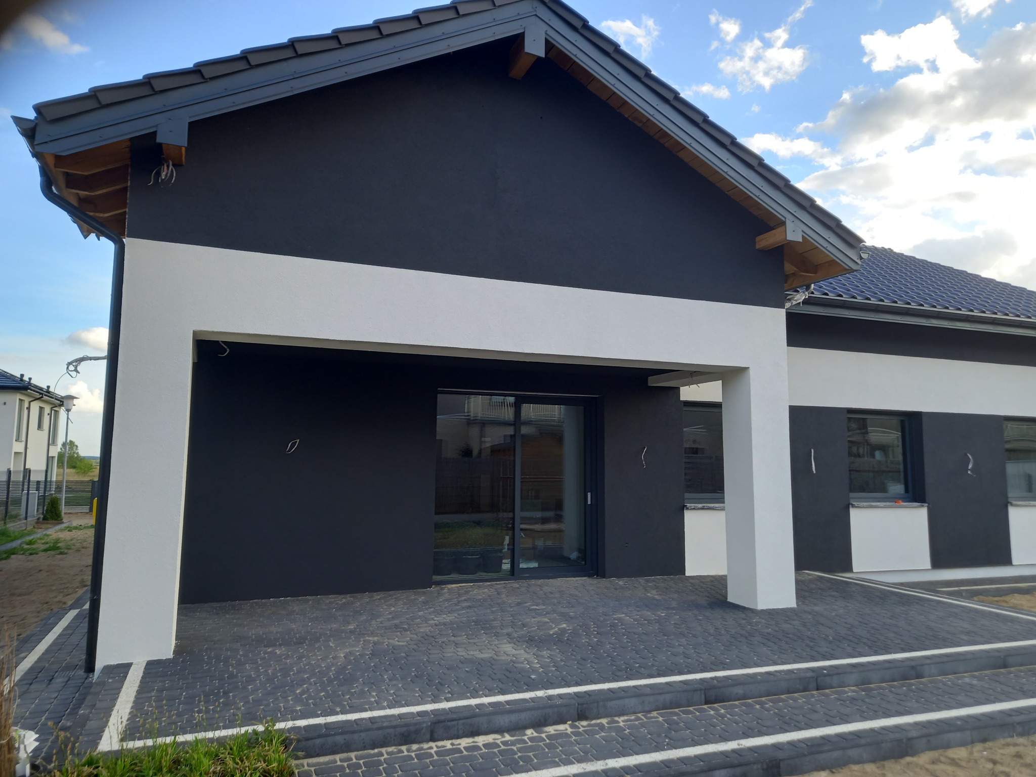 Nowoczesny dom jednorodzinny z czarno-białą elewacją, szarą dachówką i kostką brukową przed wejściem, widok z zewnątrz w słoneczny dzień.