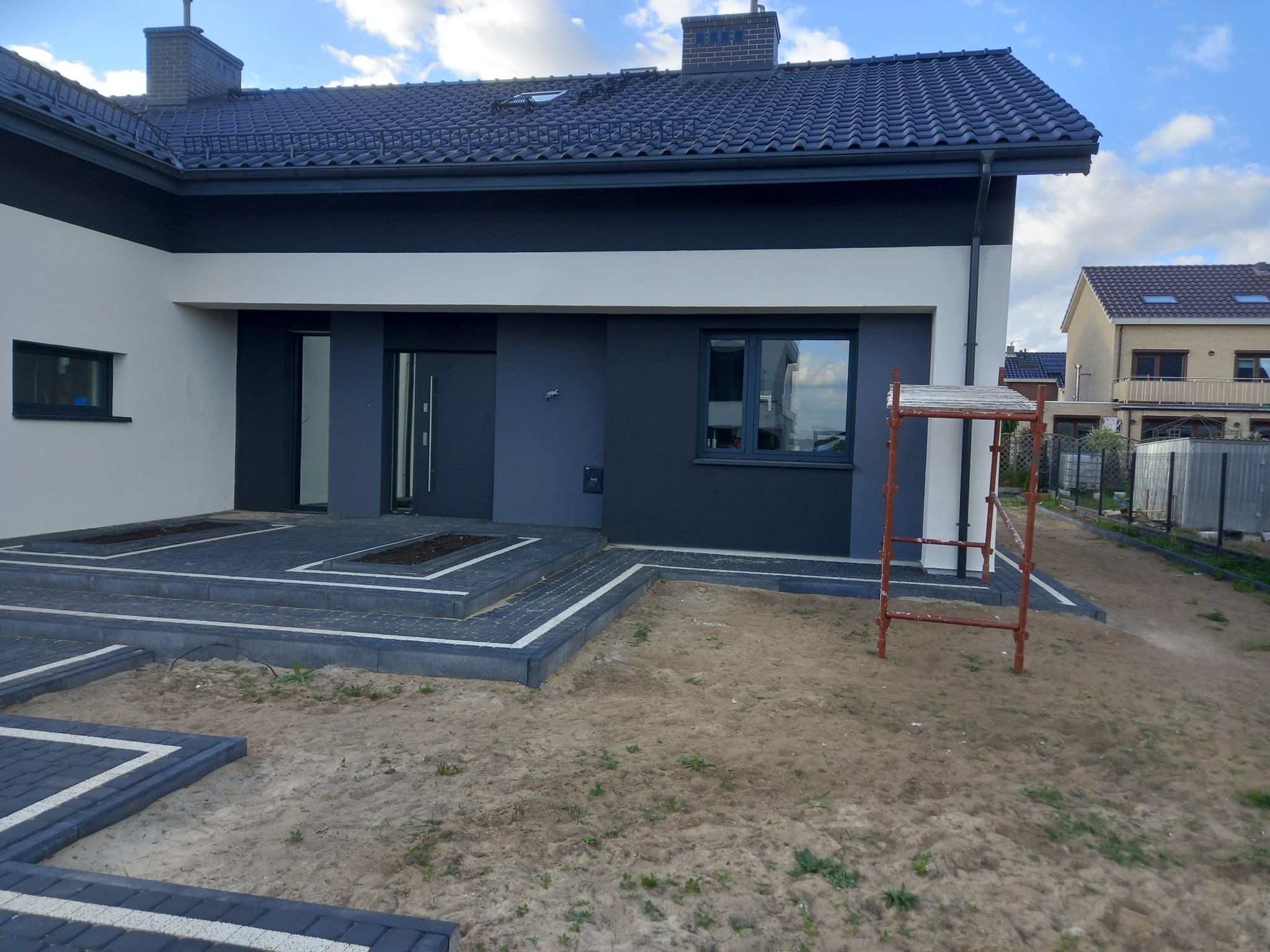 Nowoczesny dom z szarą elewacją, ciemną stolarką okienną i drzwiową oraz wybrukowanym tarasem i ścieżką z kostki brukowej w odcieniach szarości, widoczne rusztowanie malarskie.