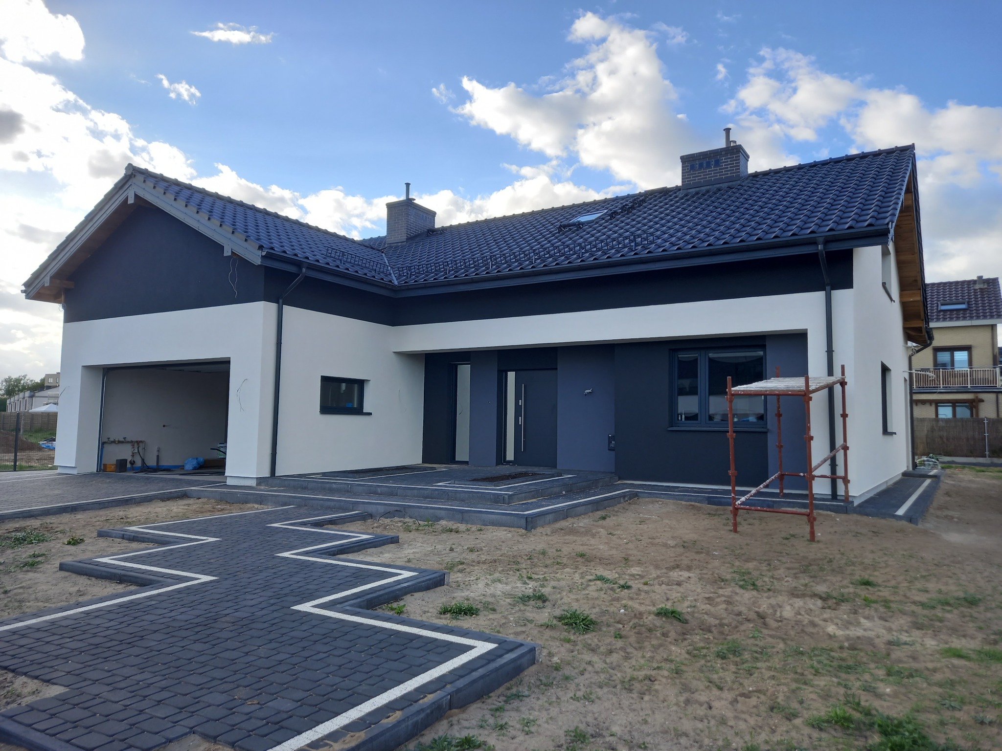 Nowoczesny, parterowy dom z dwukolorową elewacją (biało-szara), antracytową dachówką i kostką brukową ułożoną w geometryczny wzór przed wejściem, widoczne rusztowanie przy bocznej ścianie.