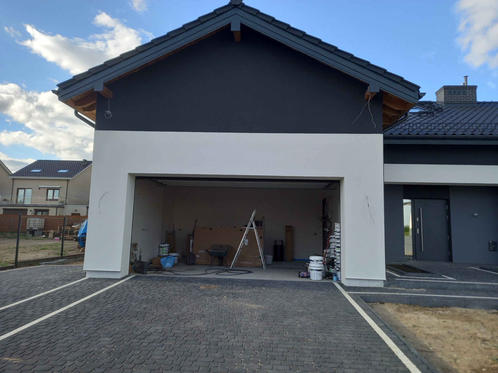 Nowoczesny dom jednorodzinny z garażem w trakcie wykończenia, widoczna elewacja pomalowana na czarno i biało, z otwartą bramą garażową, wewnątrz garażu drabina i materiały budowlane, podjazd...