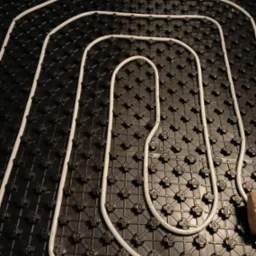 Białe rury grzewcze ułożone na czarnej płycie izolacyjnej z wypustkami, widoczne dwa cegły w rogach.