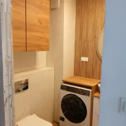 Ukończona łazienka z drewnianymi akcentami: wisząca szafka, lamelowa ściana, umywalka i pralka pod blatem, widok zza surowego otworu drzwiowego.