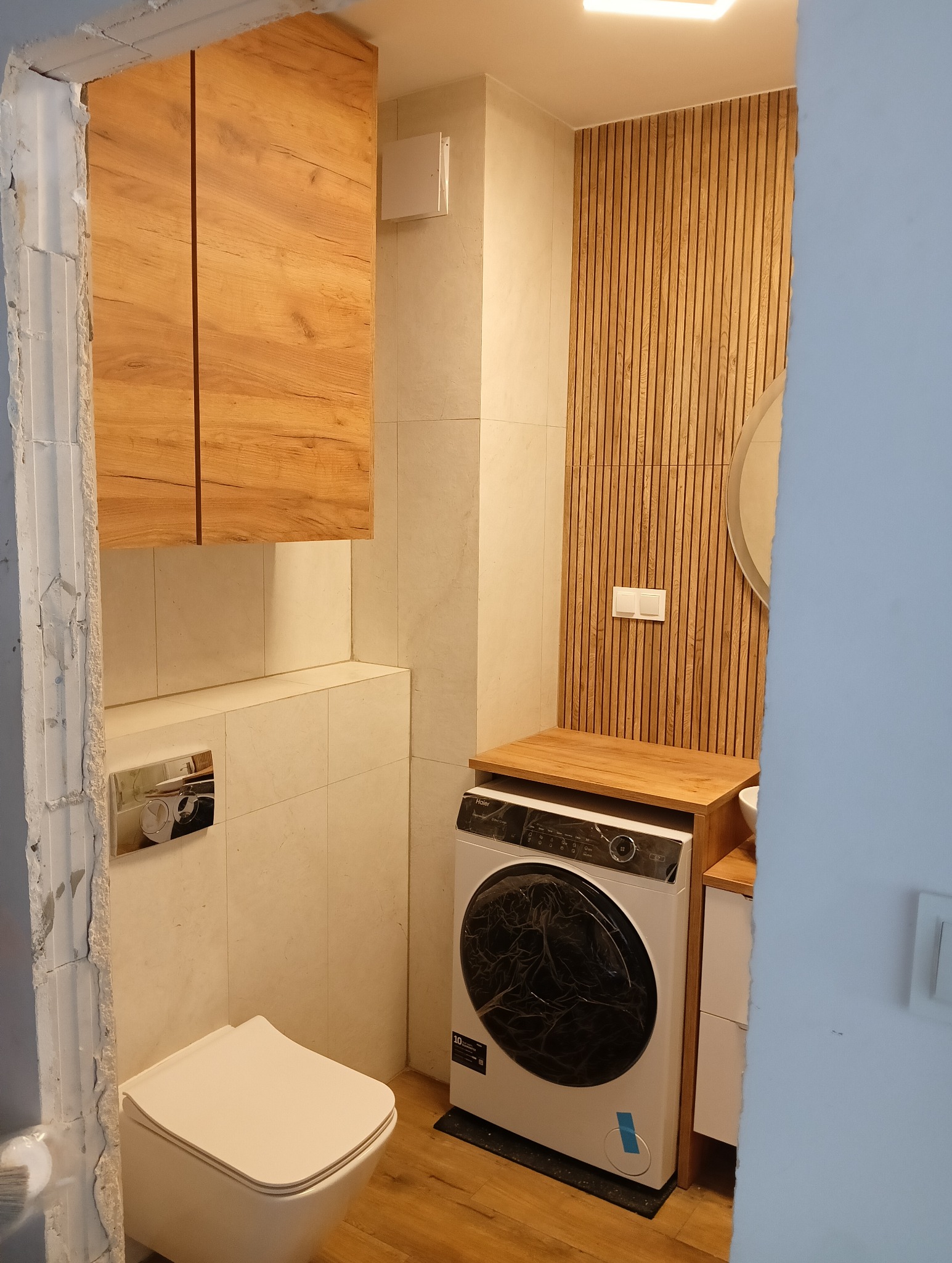 Ukończona łazienka z drewnianymi akcentami: wisząca szafka, lamelowa ściana, umywalka i pralka pod blatem, widok zza surowego otworu drzwiowego.