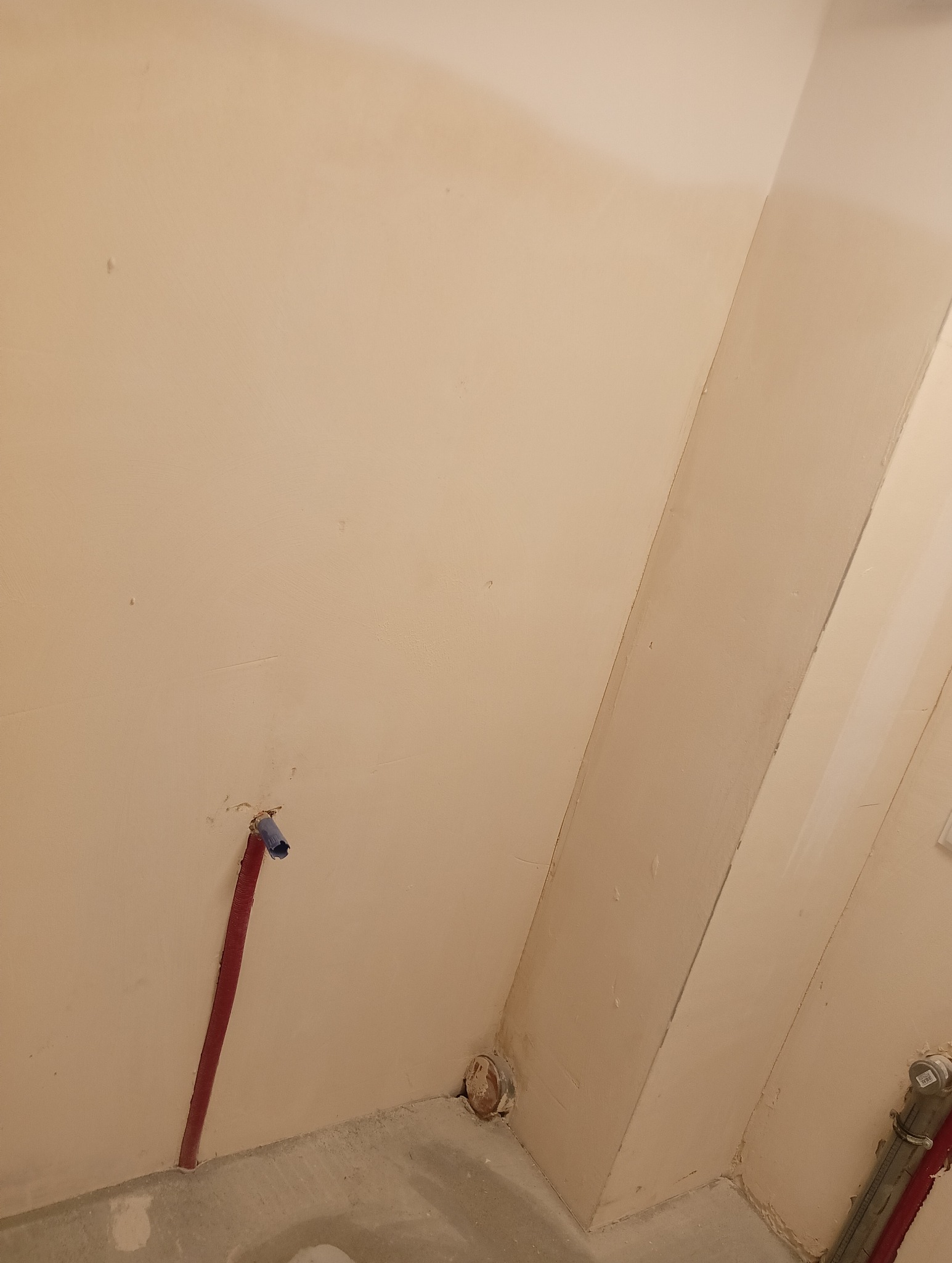 Ściana łazienki w trakcie remontu z wystającą rurą wodną w czerwonej osłonie i niebieską końcówką, oraz widoczne ślady po instalacji hydraulicznej w podłodze.