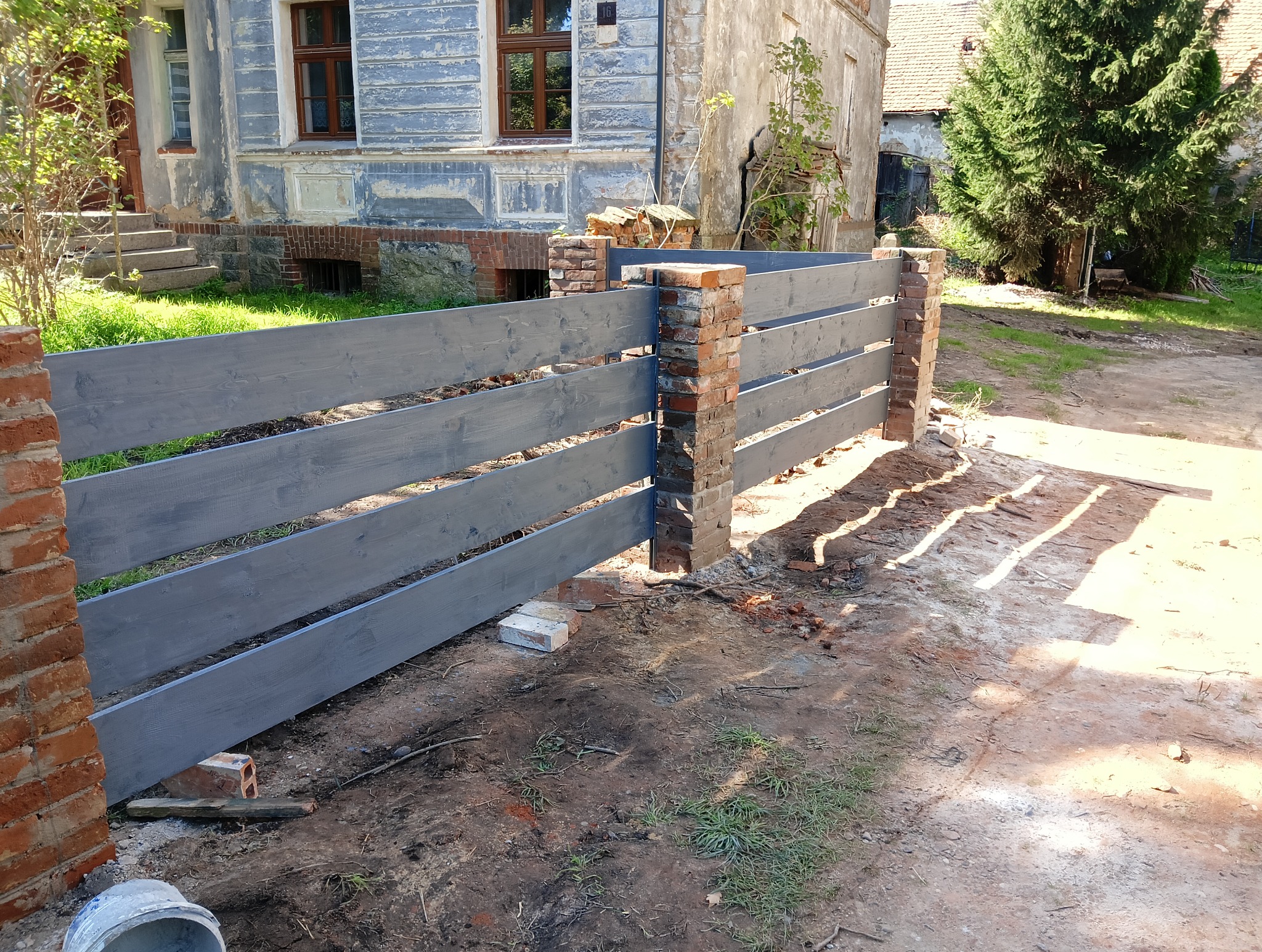 Nowo pomalowane, szare, drewniane ogrodzenie z ceglanymi słupkami na tle starego, zaniedbanego domu z łuszczącą się farbą.